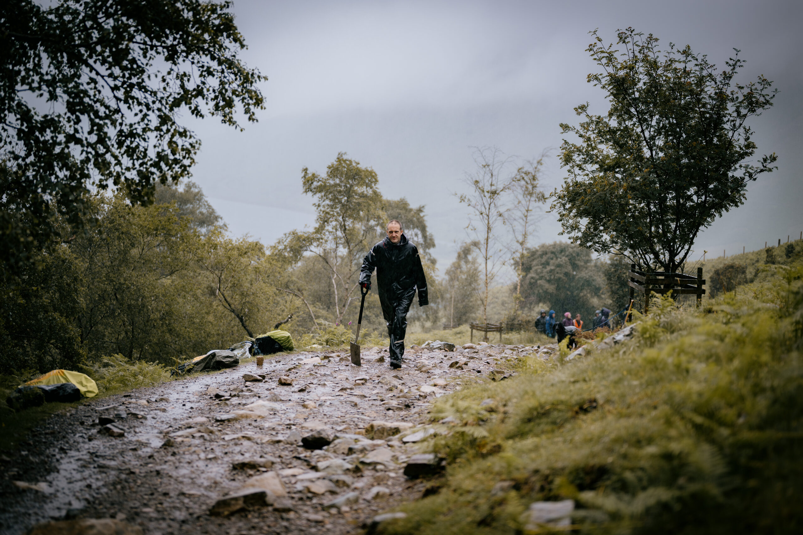 fix the fells volunteer holding a large spade walking on stone footpath surrounded by winter trees and shrubs