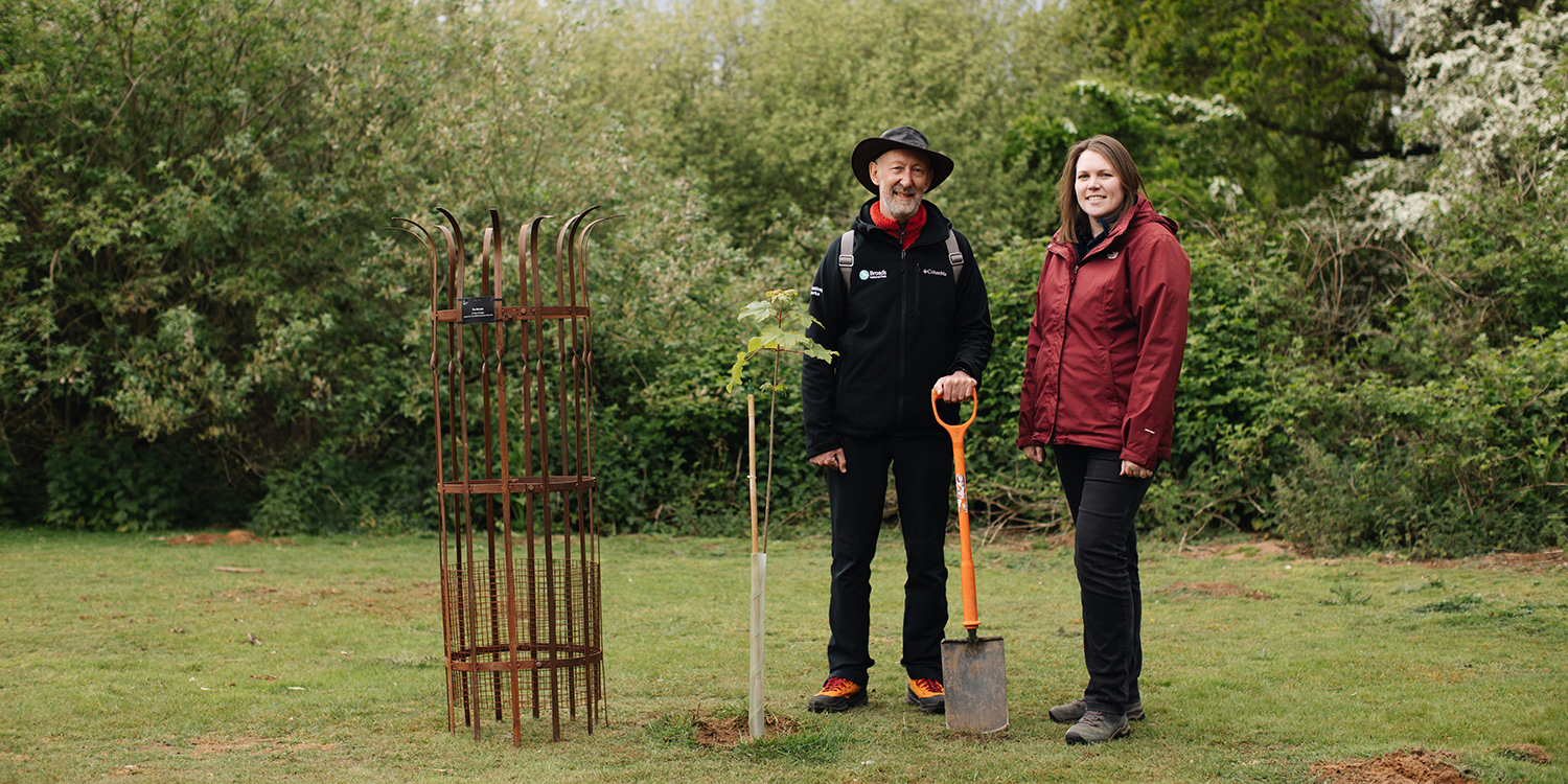 Sycamore-Gap-Sapling_John-Packman_Lynsey-Skeates.jpg Two people stand in a green outdoor setting beside a newly planted young tree and a metal tree guard; one holds a spade, with dense trees and bushes behind them.”