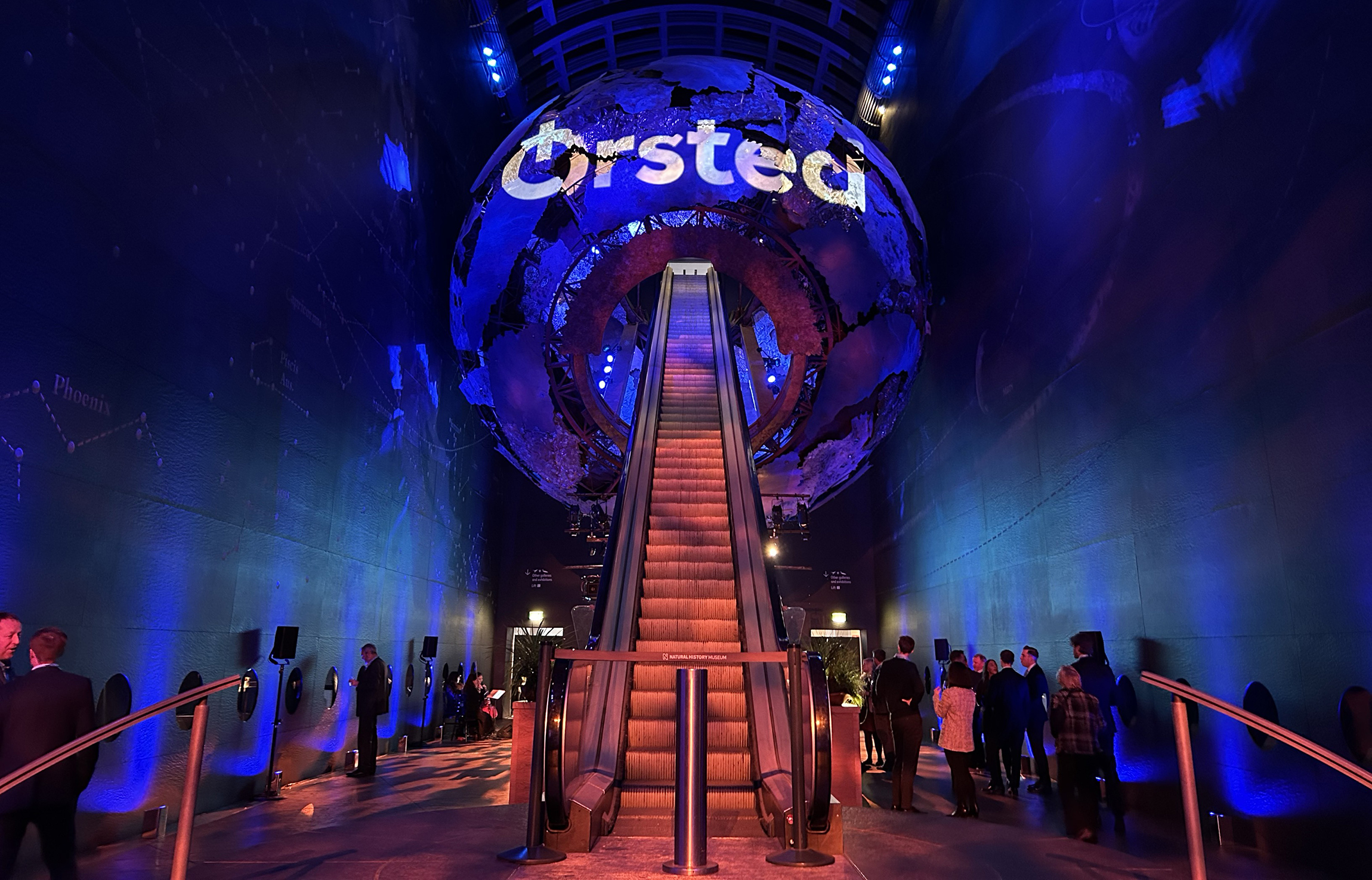 An event at the Natural History Museum in London where people mill around at the bottom of a large escalator ascending into a blue globe with the word Ørsted projected onto it