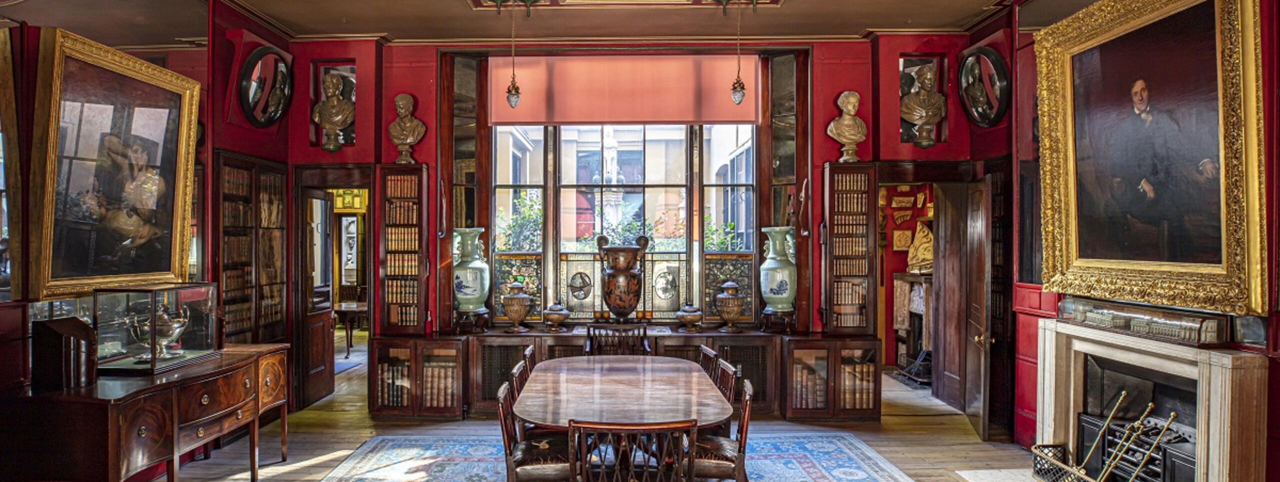 The Library-Dining Room at Sir John Soane's Museum, facing north towards a large window. On the east wall and west walls, paitings hang in gold frames. At the centre of the room, a large dining-table. By the far window, an array of antique vases.