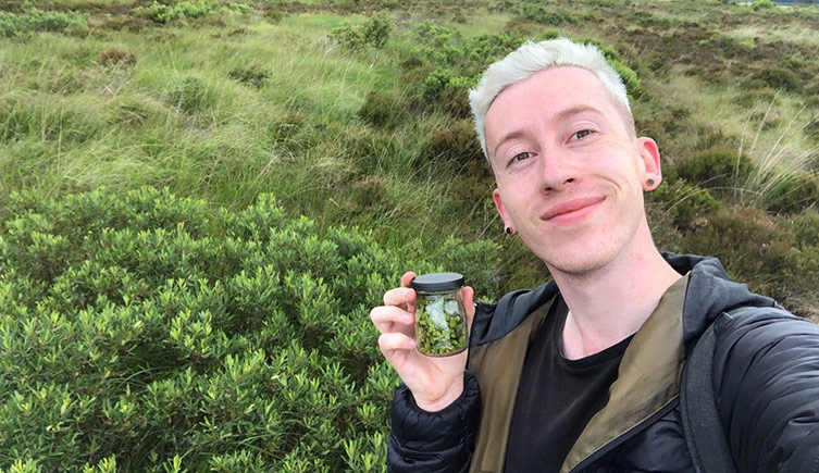 Josh Styles holding a small jar of seeds, standing in front of an extensive patch of bog myrtle on peatland