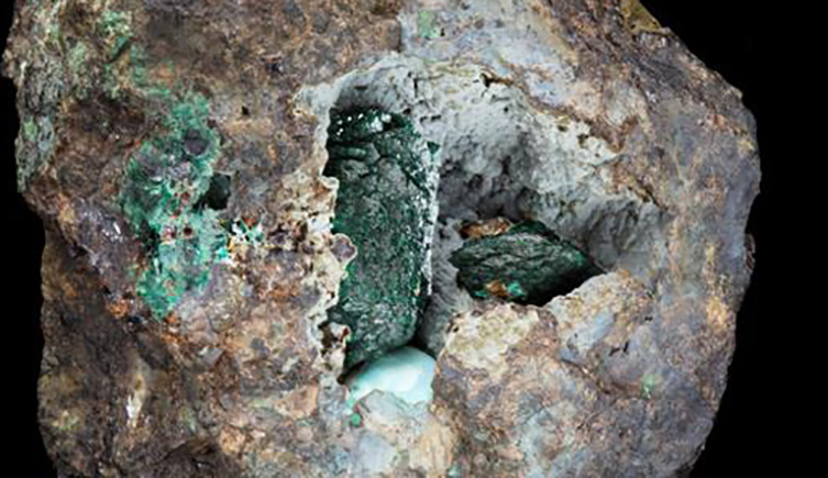 A close up showing the interior of the new mineral. Dark green crystals can be seen in the hole of a beige coloured rock, on a blackbackground.