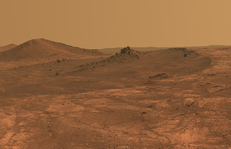 A view of Mars taken by Curiosity rover, looking out across a orange landscape with hills and valleys.