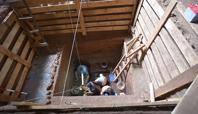 A view looking down into the excavation pit that contained the burial. The walls are supported by wood structures and four people can be seen digging at the bottom of the hole.  
