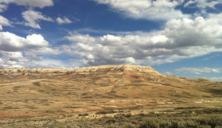 The Fossil Butte National Monument rises above a brown and green landscape beneath a blue sky filled with clouds.