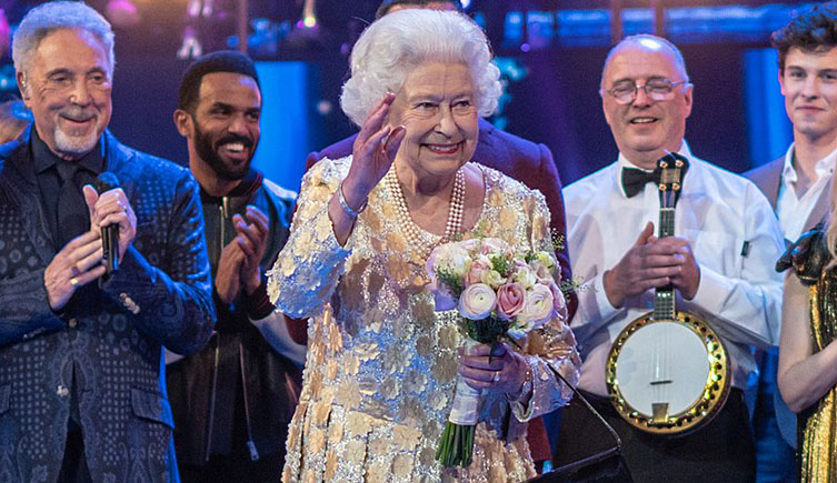 The Queen stood on the stage at the Royal Albert Hall. She is wearing a white patterned dress with pears and is holding a bunch of flowers. Behind her stands a variety of performers, including singers Sir Tom Jones and Craig David.
