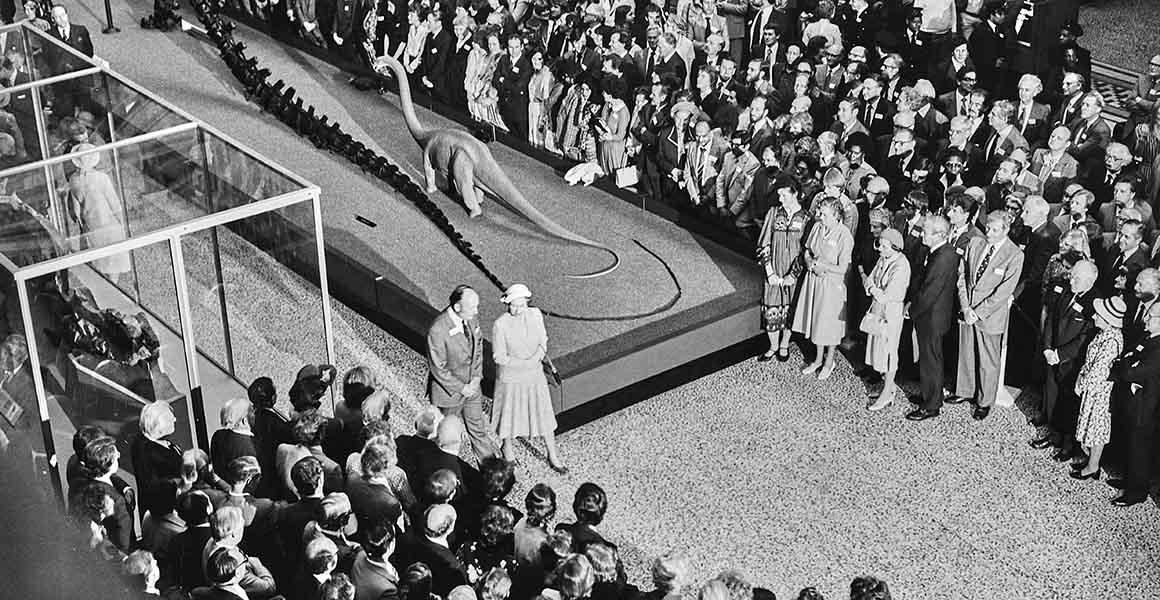 A black and white photograph of The Queen walking through Hintze Hall, surrounded by a large crowd of onlookers. Behind her, the tail of Dippy the Diplodocus is visible.
