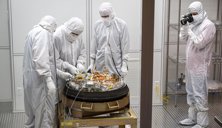 The team at NASA opening the capsule containing the samples. Three people are stood around wearing white overalls. 