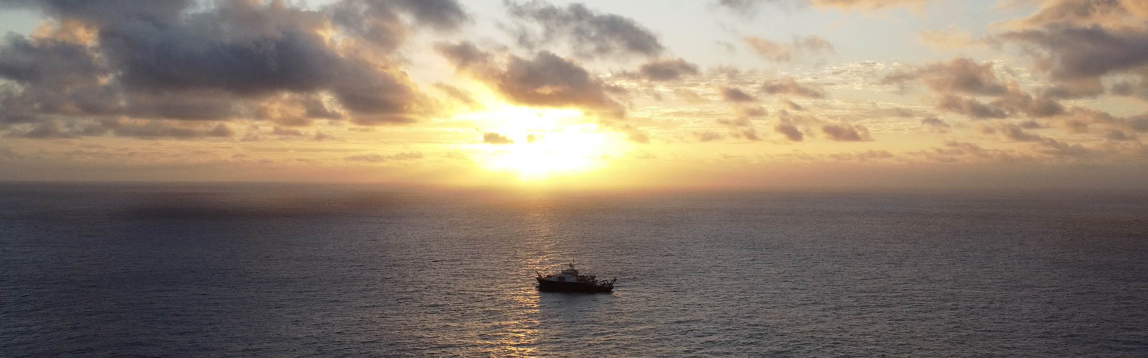 A picture looking out across the vast Pacific Ocean as the dun sets over the horizon. In the middle of the water is the silouette of a boat.