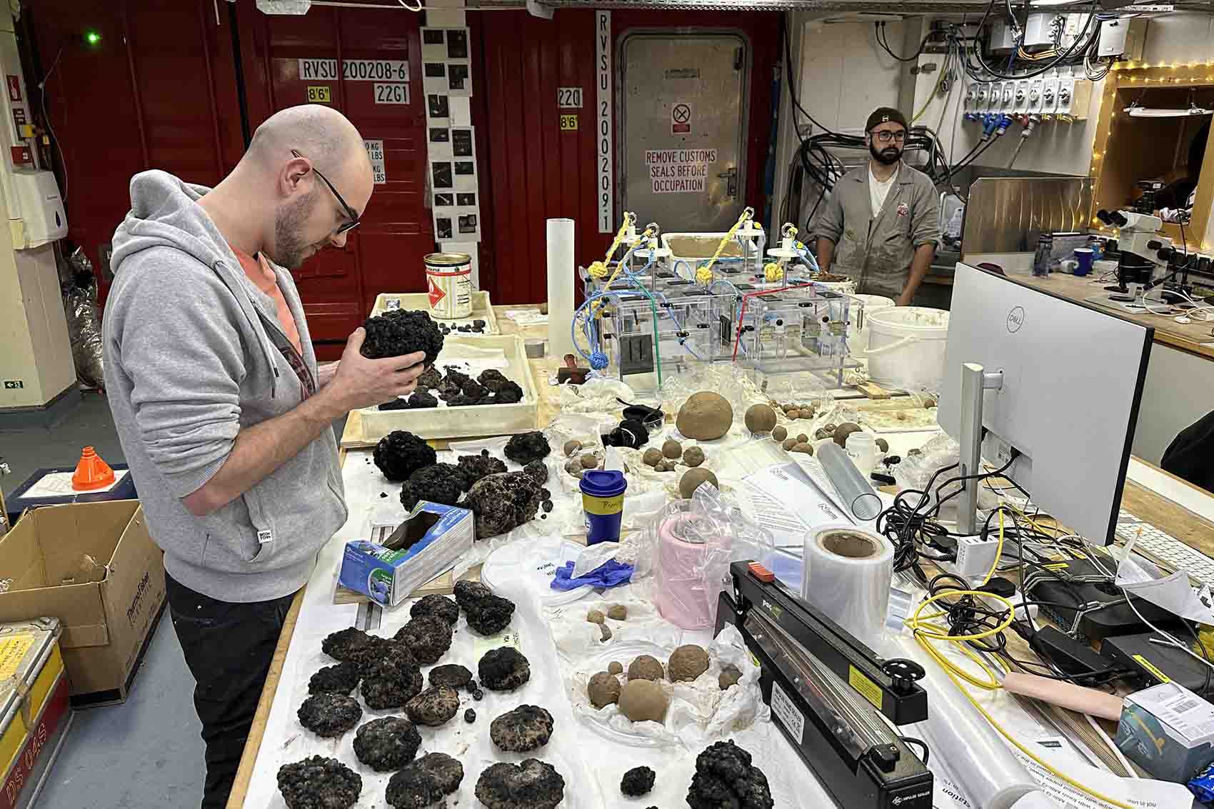 A picture taken from within a lab on a research boat. A person stands at a table holidng a large nodule, while the table in front is covered with more nodules and other pieces of scientific equipment.