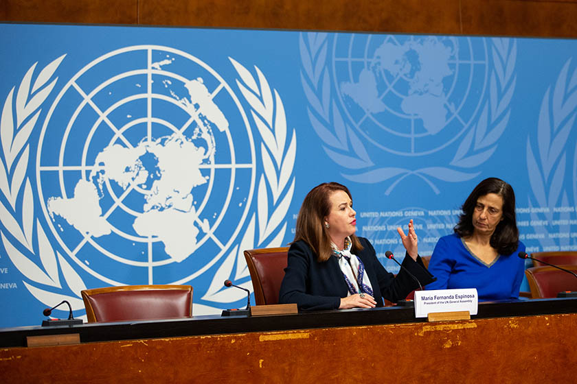 A picture taken from a a United Nations conference, with two people sat behind a desk in front of the blue and white UN logo.