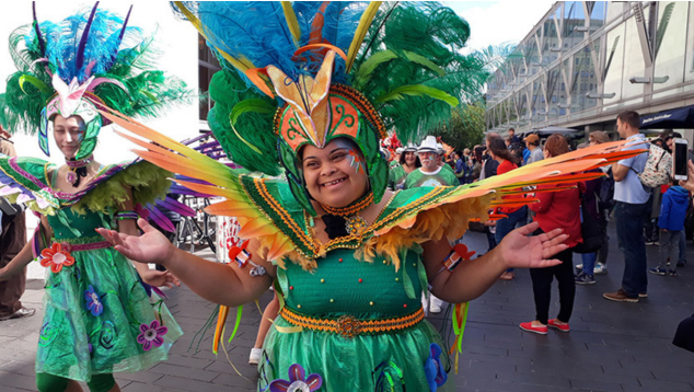 A woman stands with her hands facing outwards. She’s wearing a bright carnival costume with a large headdress of feathers. She’s standing among a parade of people, similarly dressed. Viva Carnival Club, Alegria Samba School. Photo by Paul Davies