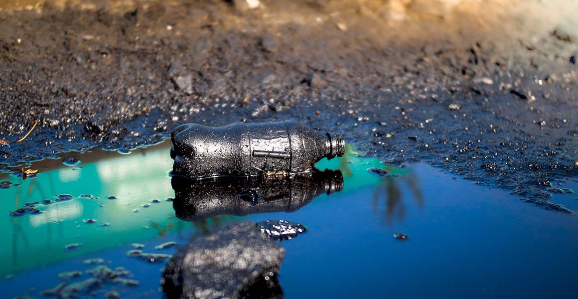 A plastic bottle stained black lies in a pool of black liquid on bare soil.
