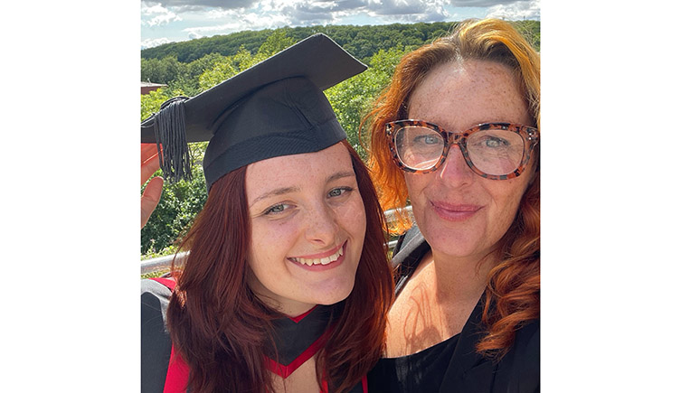 Daisy Cadet smiles broadly at the camera while wearing a graduation cap, next to her mother Ashleigh who is wearing large, thick rimmed glasses.