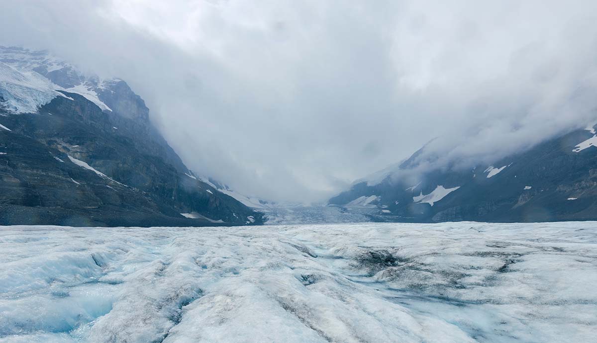 Mountains shrouded in cloud rise above the ice sheet of the Athabasca Glacier.