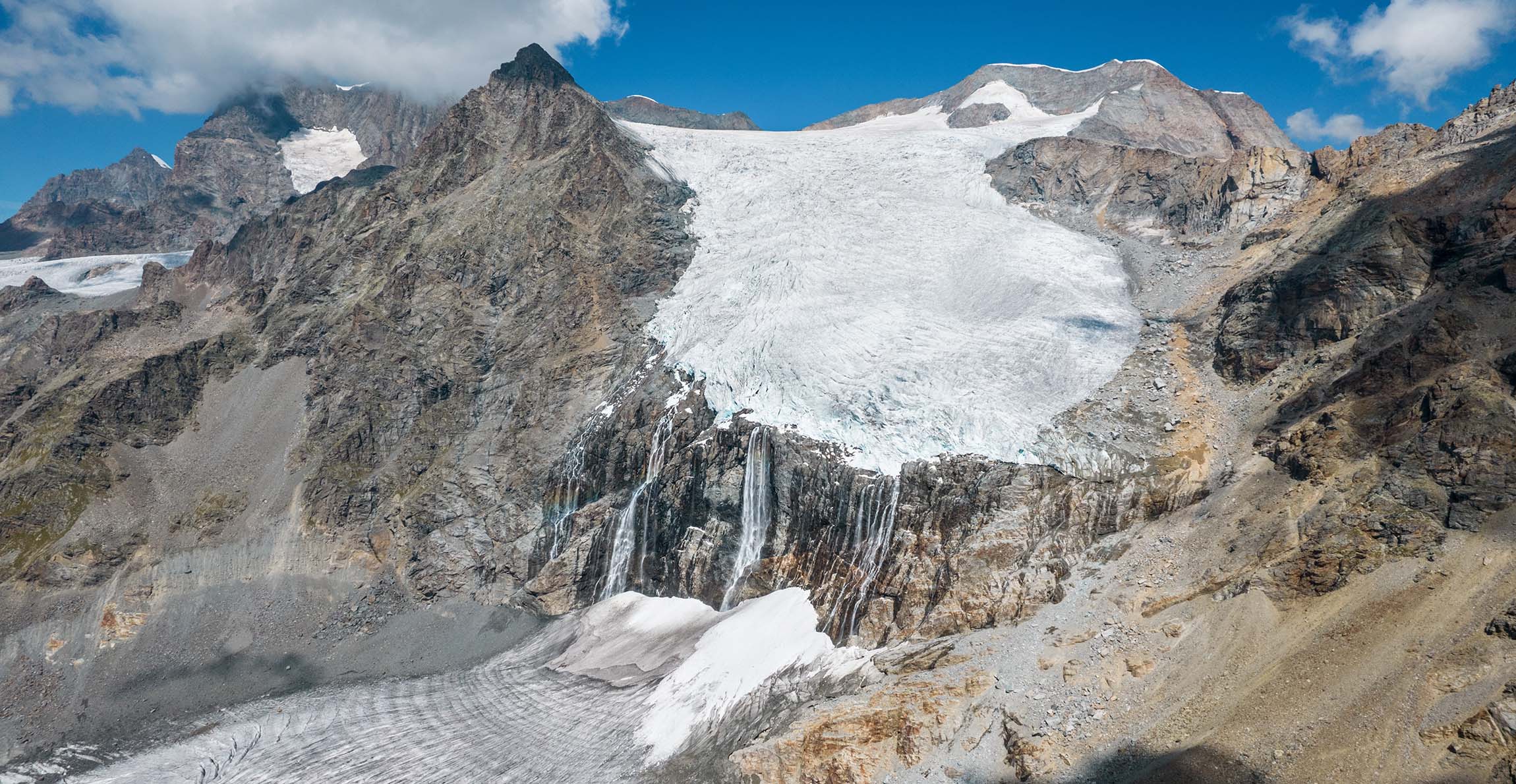 A photo of the Fellaria glacier in Italy, with meltwater flowing down the mountain from a large icy mass sandwiched between two peaks.