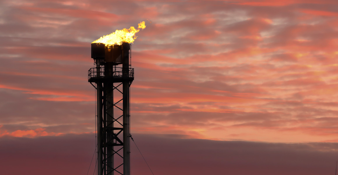 Fire burns atop an industrial chimney against a red evening sky.