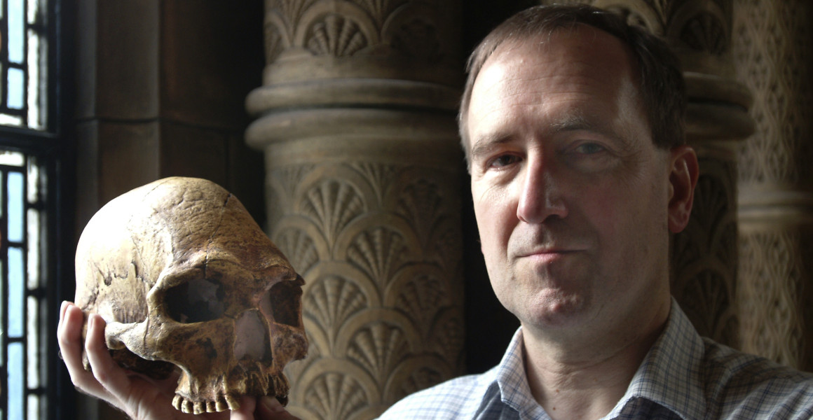 Professor Chris Stringer holds a hominin skull near to a window with an intricately decorated column behind.