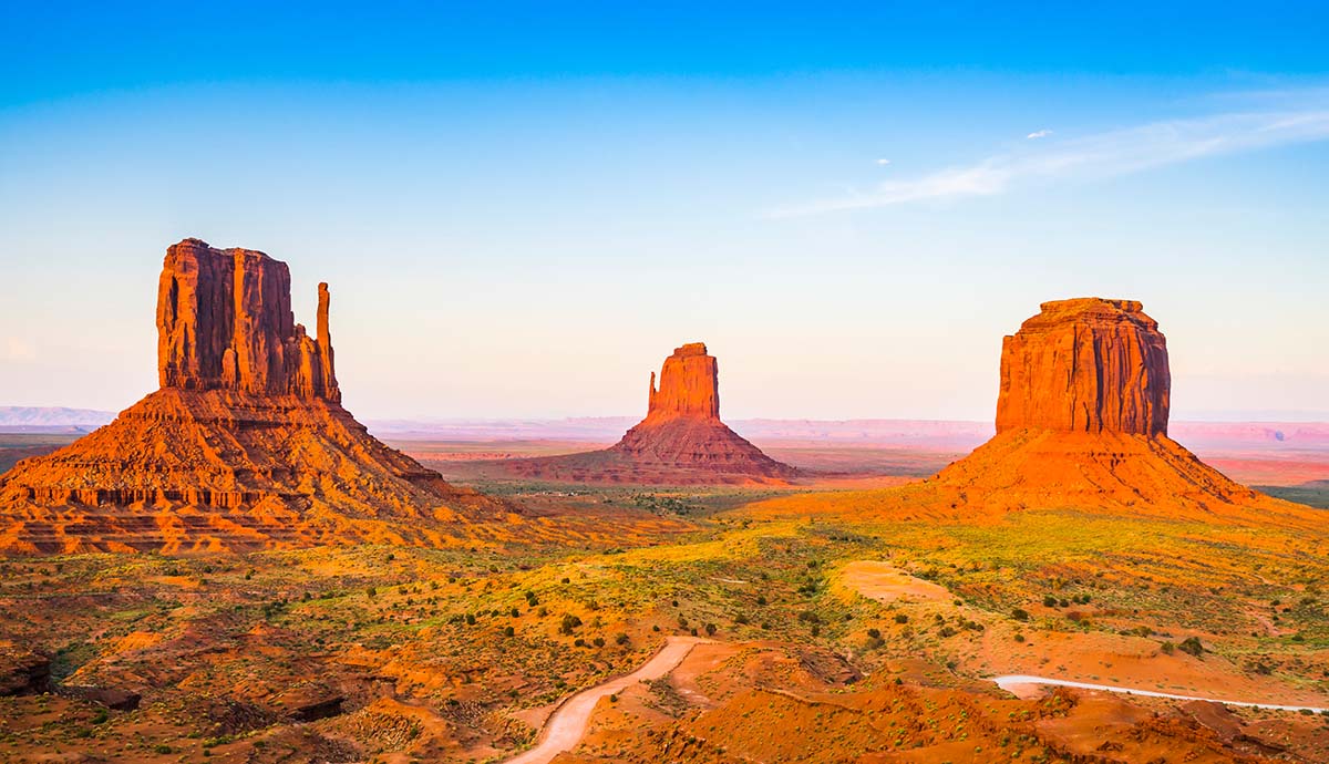 A photo showing three sheer-sided rock faces rising up above an orange landscape covered in shrubs in Monument Valley.