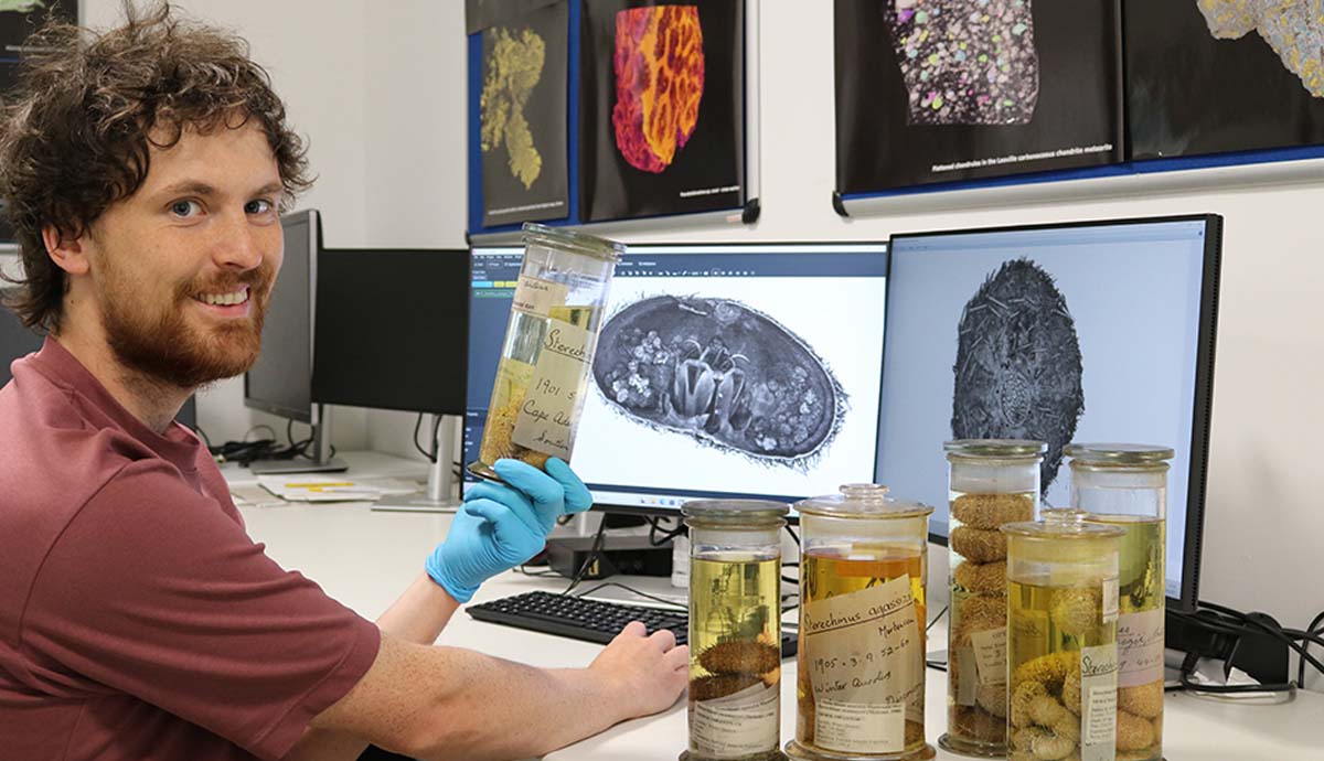 Hugh Carter sitting in front of a computer. He is holding a jar containing a specimen, while looking over his shoulder at the camera and smiling. 