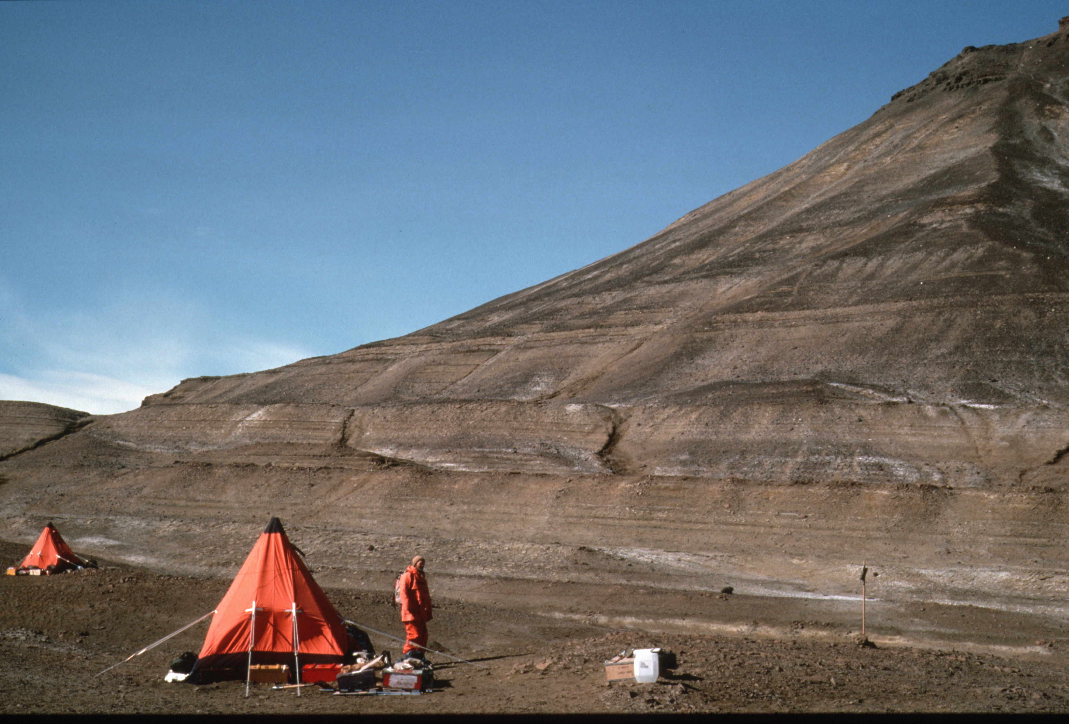 A rockey grey landscape, broken up by bright red reds in the foreground. Next to one of the tents stands the figure of a person in a red coat.