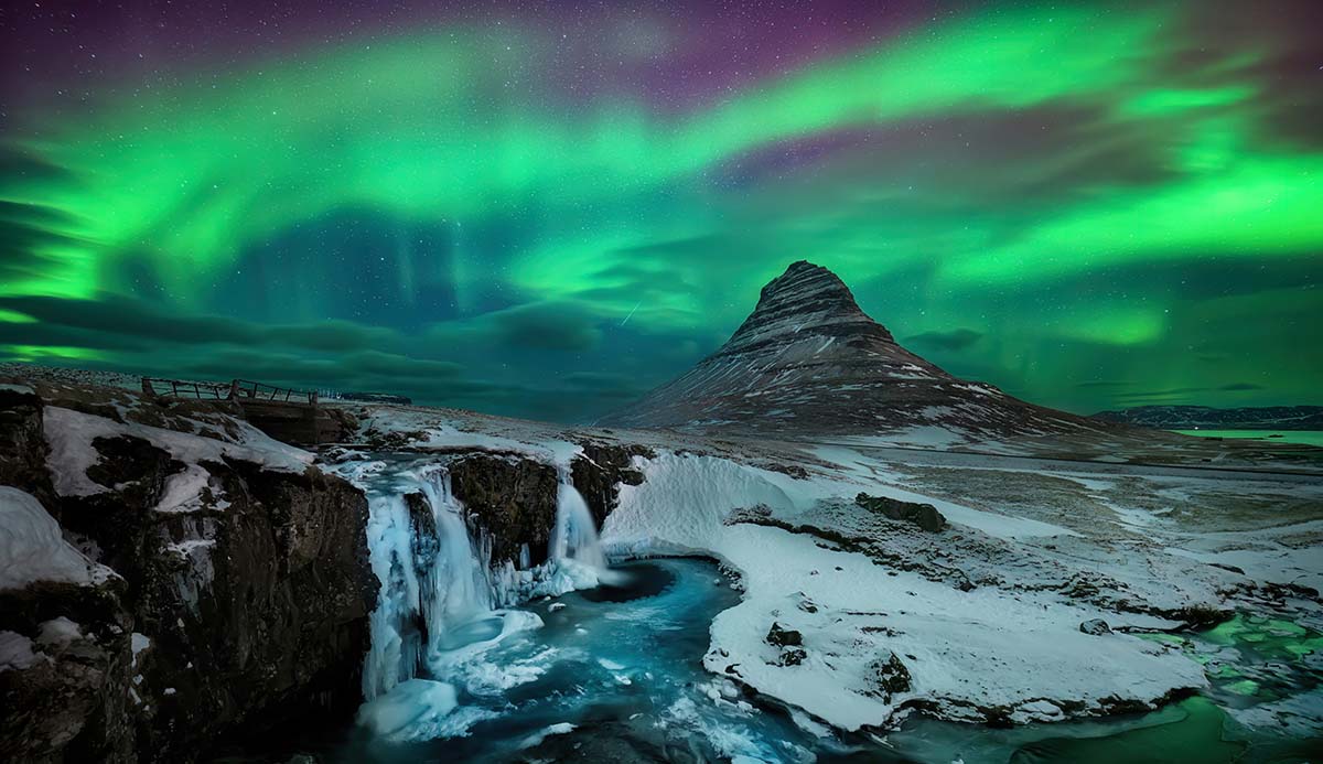 A green aurora hangs over Kirkjufell mountain in Iceland, with snow-covered ground and a waterfall in the foreground.