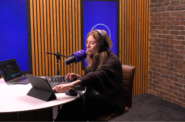 Lady in radio studio with headphones on, microphone in front of her, at a desk, two laptops next to her, blue screens behind her.
