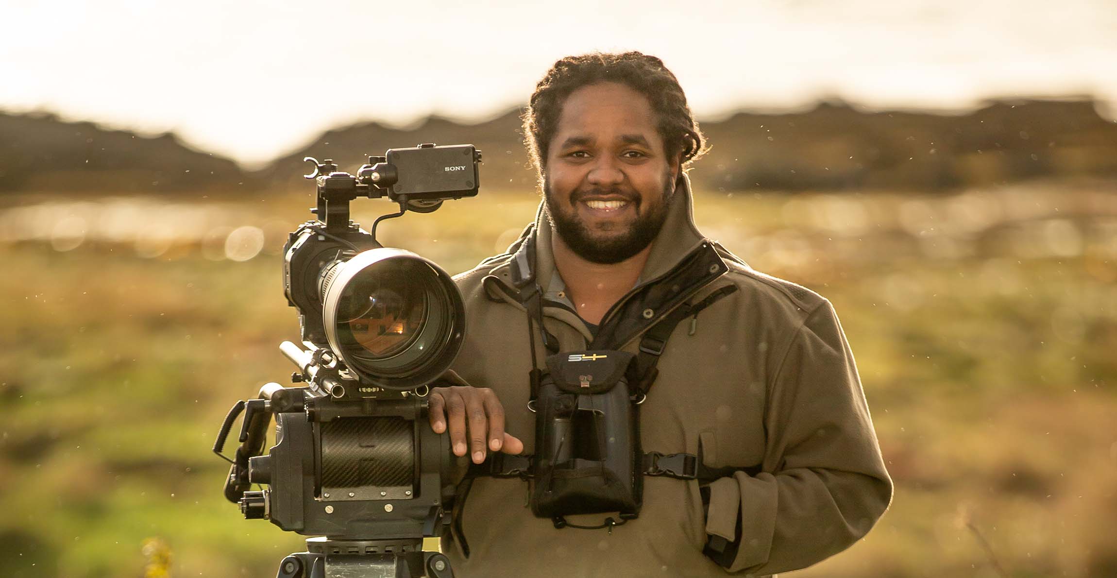 Hamza Yassin, who has a pair of binoculars around his neck, smiles as he stands with a hand resting on a video camera tripod. 