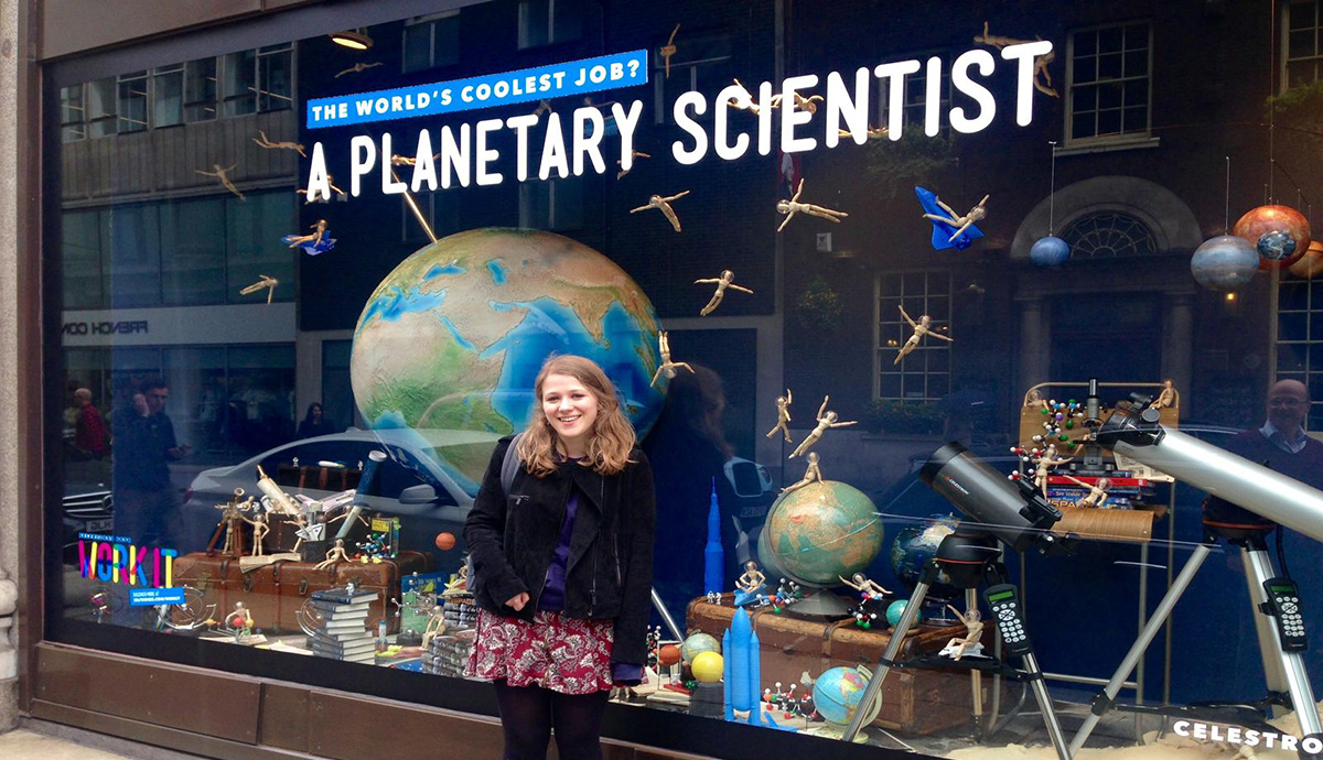 Dr Helena Bates in front of a shop window display with text that reads: "The word’s coolest job? A planetary scientist."