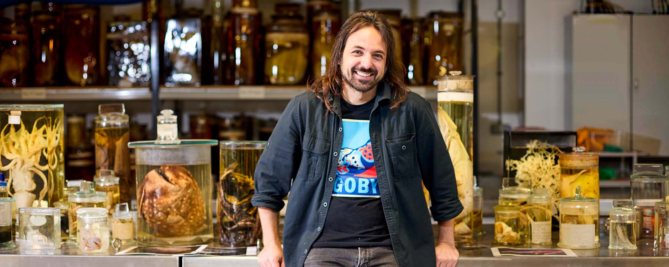 A photo showing Dr Diego Vaz, Senior Curator of Fish, leaning against a metal container in our Tank Room. A variety of glass jars containing specimens of fish and other marine life are behind him.