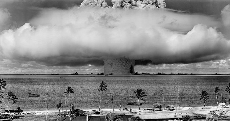 Black and white image of a nuclear weapons test over water. You can see an inhabited beach in the foreground.