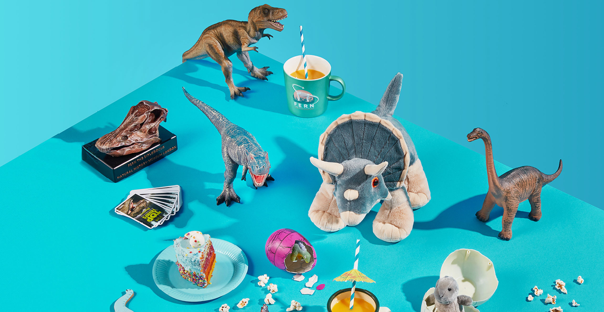Dinosaur-themed toys in a party setting against a blue background. The toys include T. rex models, a Diplodocus replica skull and a cuddly toy Triceratops.