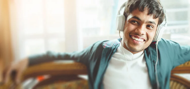Asian, Indian happy young man listening podcast through headphones. He is sitting relaxed on couch near window at day time.