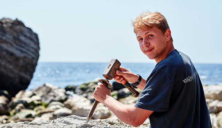 A photo of Jack Wonfor using a hammer and chisel to extract a fossil from a boulder