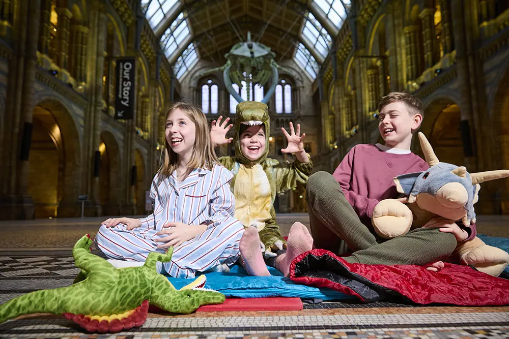 A family of a mother, a father and three children sit in their pijamas on sleepig bags under Hope the blue whale, in Hintze Hall