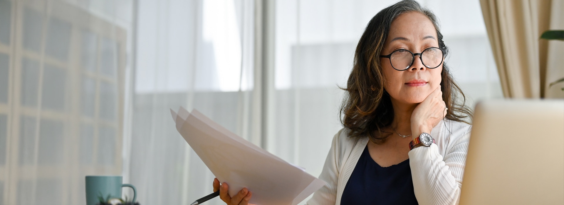 A woman holding a stack of papers looks at a a laptop screen