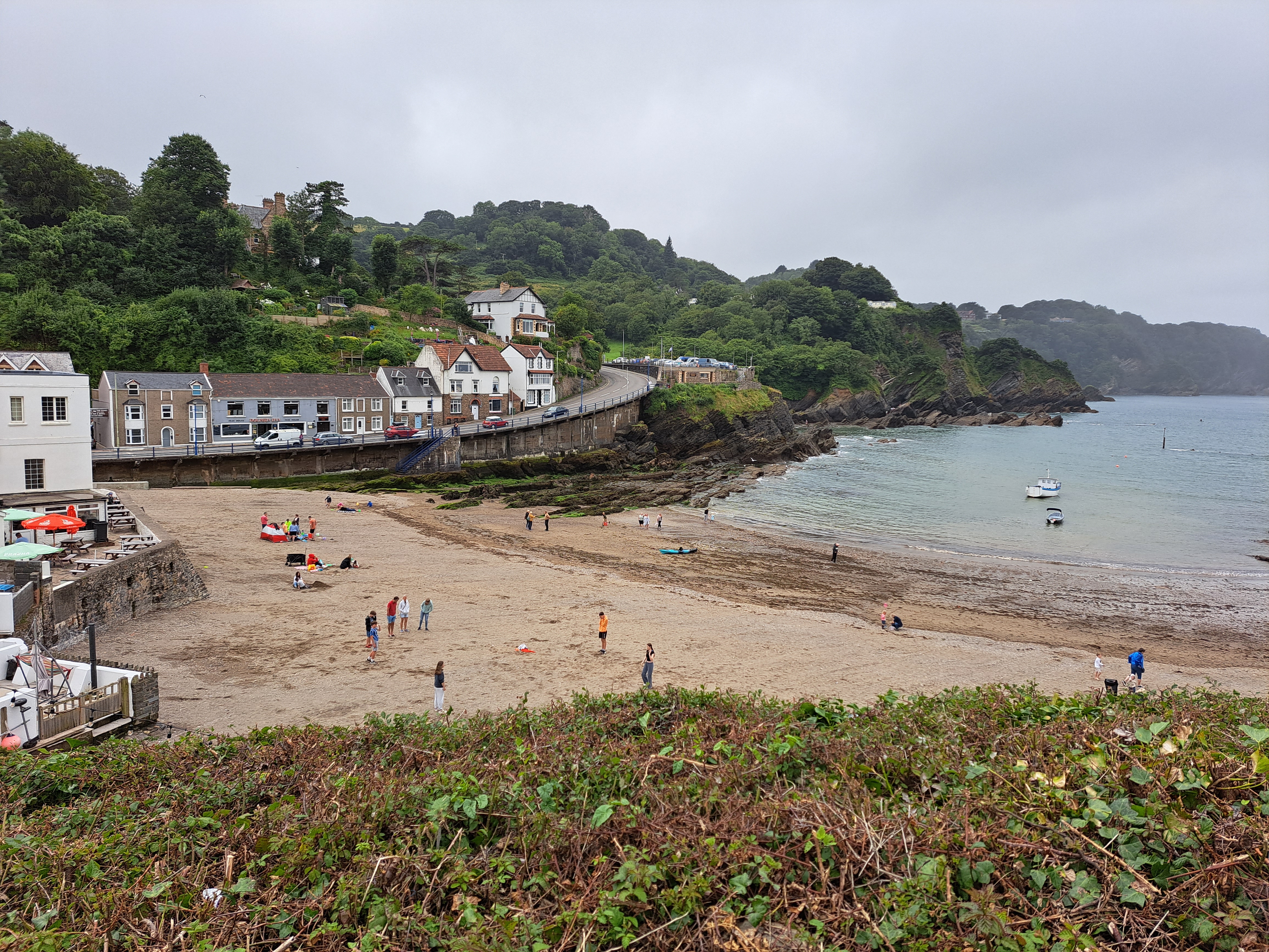a beach with people on it in front of cottages and the sea beyond which has boats in it