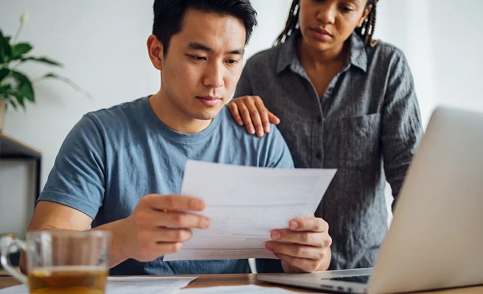 A man studies a sheet of paper. A woman stands behind him with her hand on his shoulder.