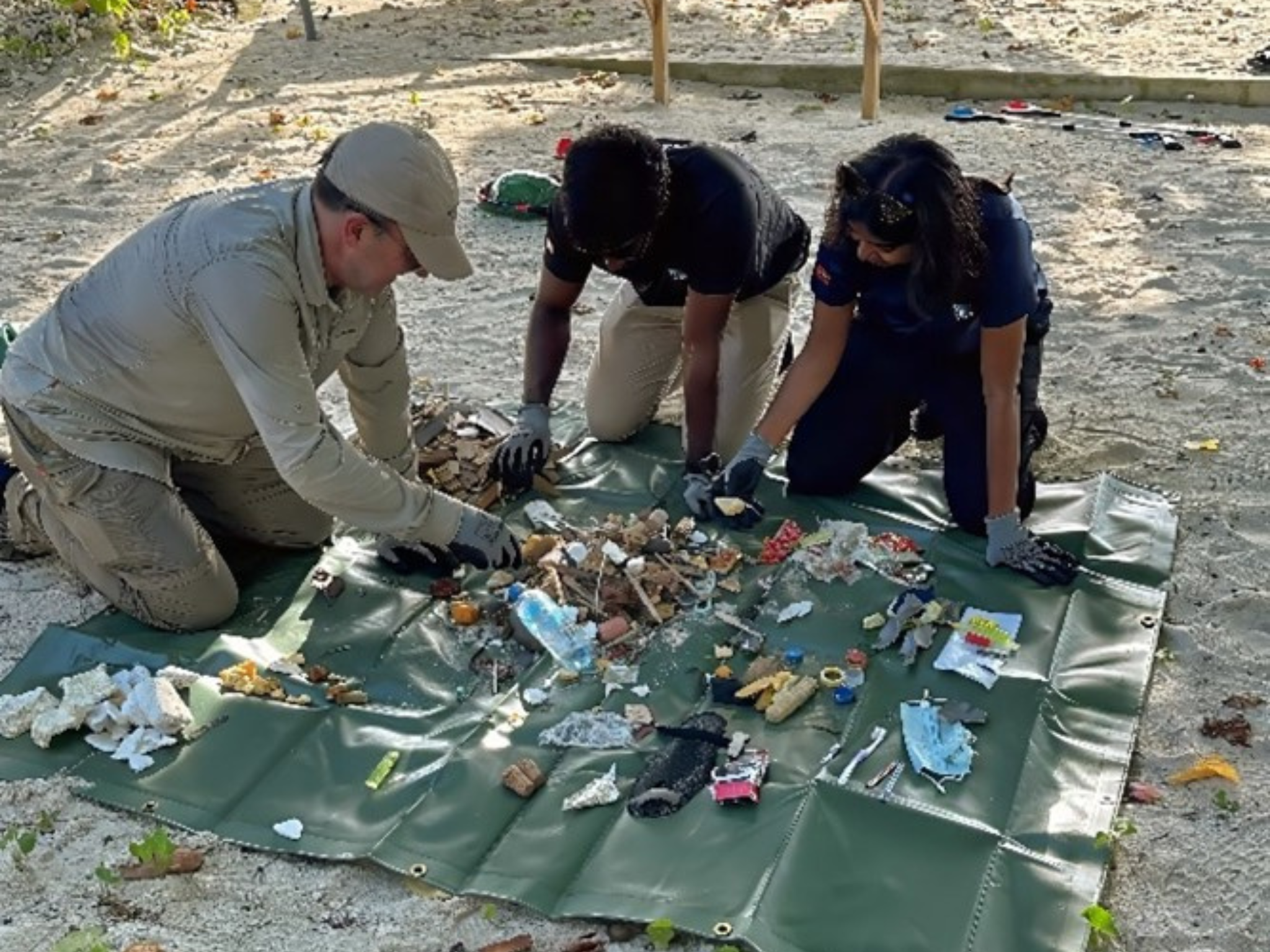 three people on their hands and knees leaning over a piece of tarp, sorting through a pile of various plastic pieces and litter