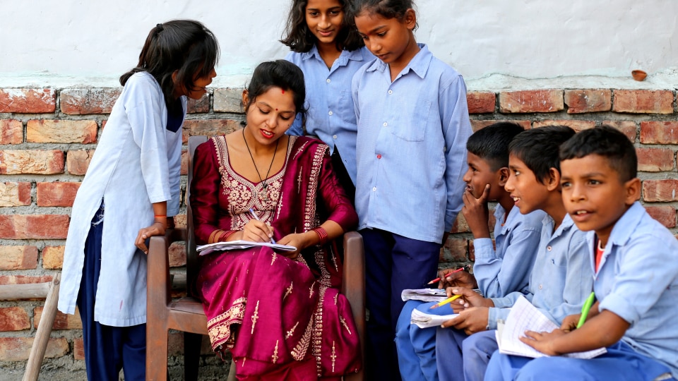 A South Asian teacher sitting outside surrounded by her learners