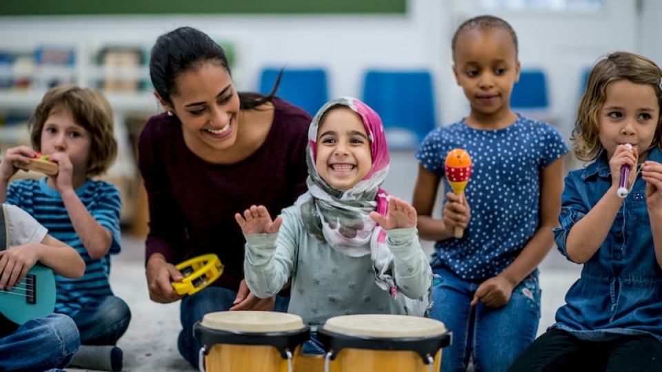 Five young learners of mixed ethnicity playing various music instruments. Their teacher kneels besides them, smiling