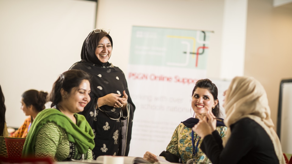 A group of women teachers in a classroom, sitting and talking together while one teacher stands beside a whiteboard.