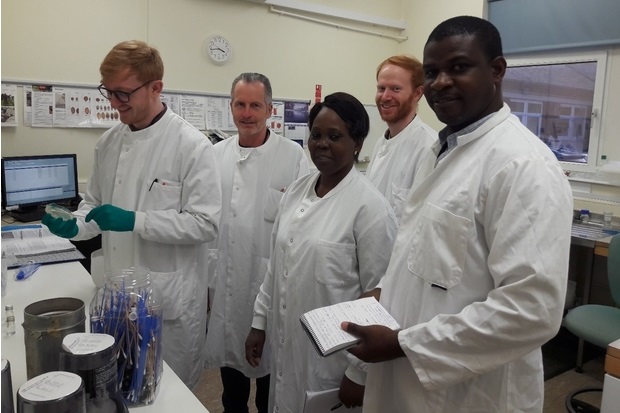 Five diverse laboratory scientists in white coats pose in a clinical lab; one holds a notebook, while test tubes and equipment surround them.