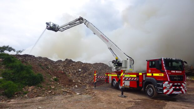 This image shows a fire truck and crew attending a waste fire. The crew are using a crane at the back of the truck to extend a platform over the waste fire to enable the fire officer to spray the waste fire from above.