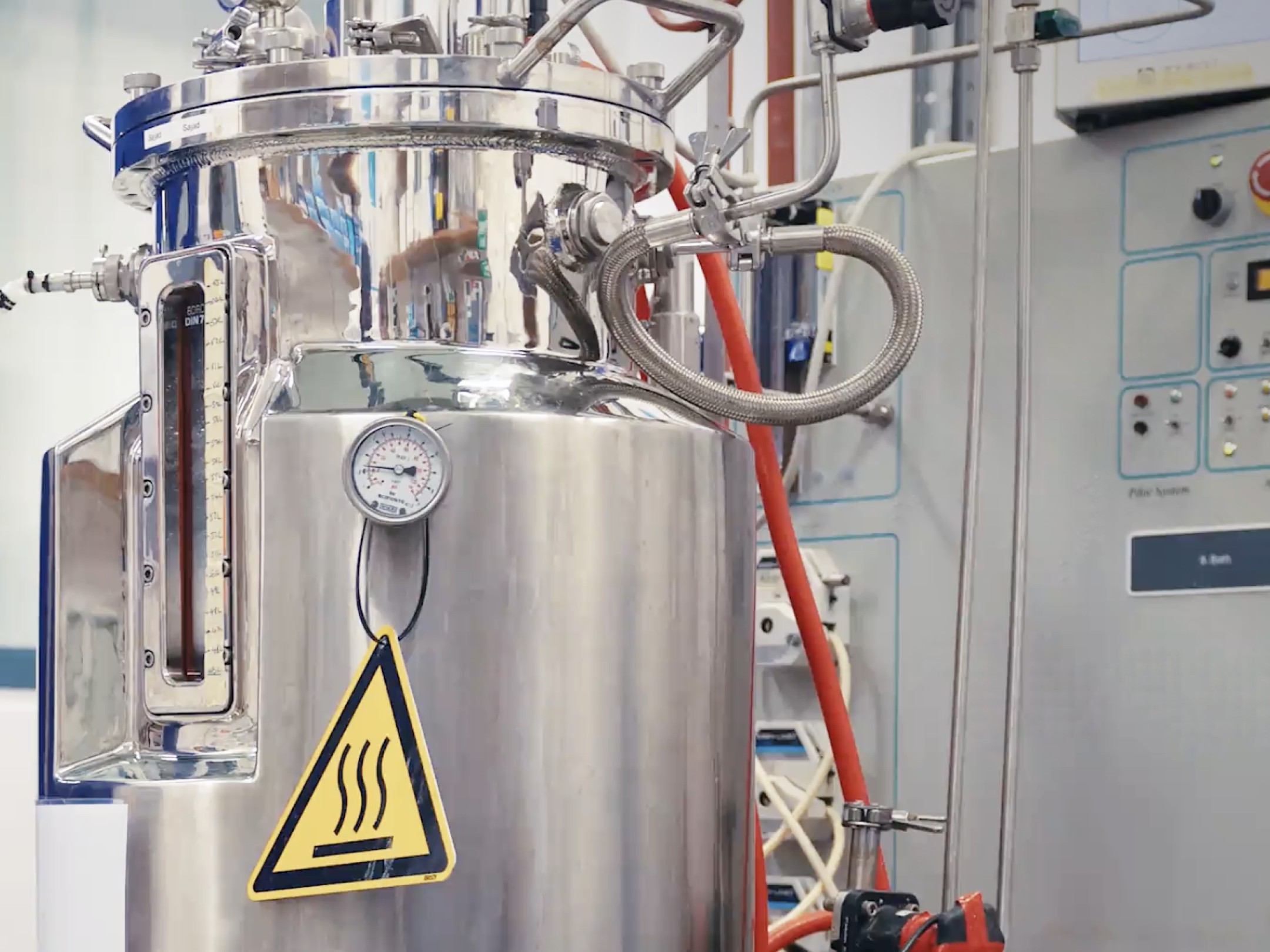 Stainless steel fermentation tank with pressure gauge, pipes and heat warning sign in a lab setting.