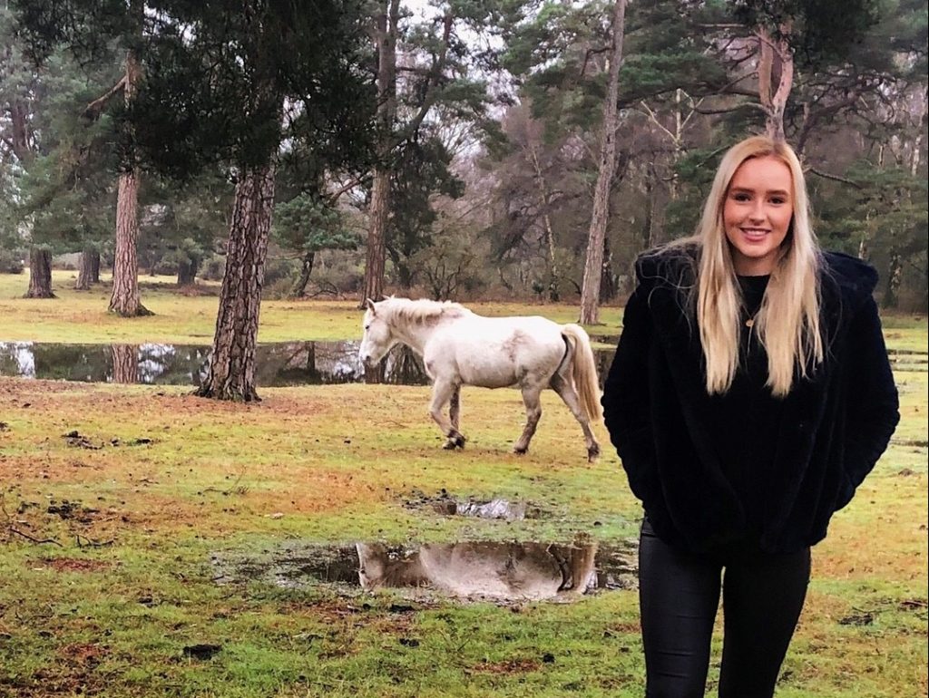 Picture of Rebecca in a black fur coat and jeans. She has blonde hair and is standing in a woodlands with a white horse behind her.