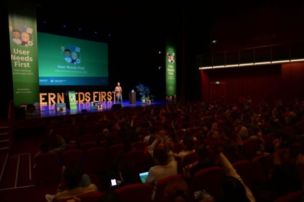 The MC standing on the stage of the User Needs First Conference. The auditorium is dark and the audience is just about visible. The stage has ceiling to stage banners and a slide projected - all with the User Needs First Community green branding.