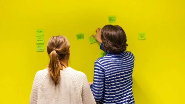 Photograph of two people sticking green post its with writing onto a bright yellow wall. The people appear to be in discussion.