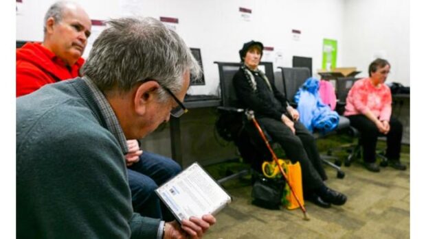 People sat in a waiting room in a library. A man with grey hair and glasses facing away holding a CD. A chap in a red jumper sits waiting, along with a lady wearing black and man with a pink jumper.