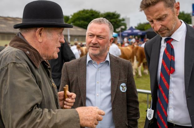 Steve Reed, Environment Secretary, stands outdoors at a livestock show talking with two men. He is in the centre, wearing a brown jacket and light blue shirt, listening. On his left, an older man in a dark coat and black bowler hat gestures while speaking. On his right, a suited man with a red and blue striped tie looks down thoughtfully. Cattle and people are visible in the background.